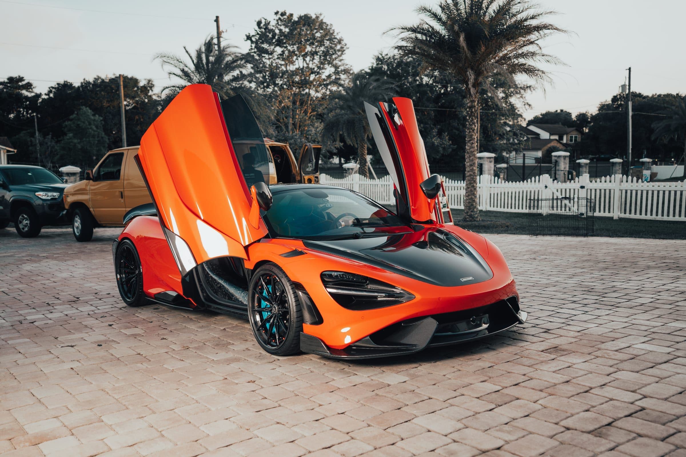 Orange McLaren with both dihedral doors raised, parked on a paver driveway with palm trees and a white picket fence behind it